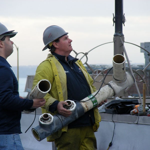 Two Performance Mechanical Group’s technicians holding a complex pipe structure on city rooftop.