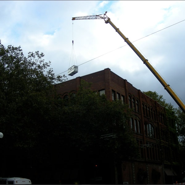 Crane lifting a large piece of equipment or a large container, over a brick building.